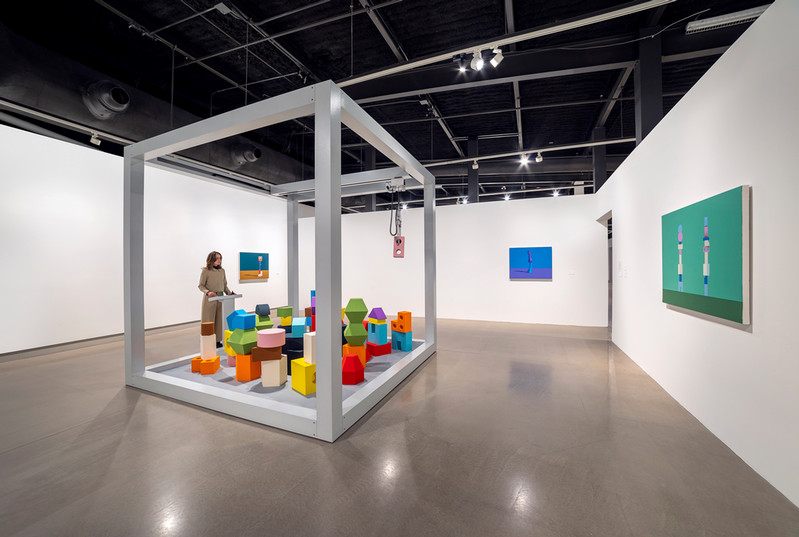 Jennifer Marman and Daniel Borins, Balancing Act, 2023. Image from Contemporary Calgary. (An art installation that consists of a room-sized claw game with colourful shapes, and paintings on the wall with figures constructed from the same shapes)